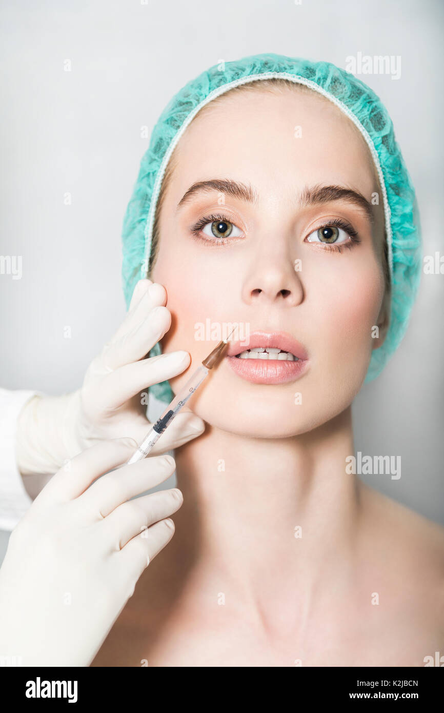 Doctor aesthetician makes hyaluronic acid beauty injections in the nasolabial fold of female patient in a green medical cap Stock Photo