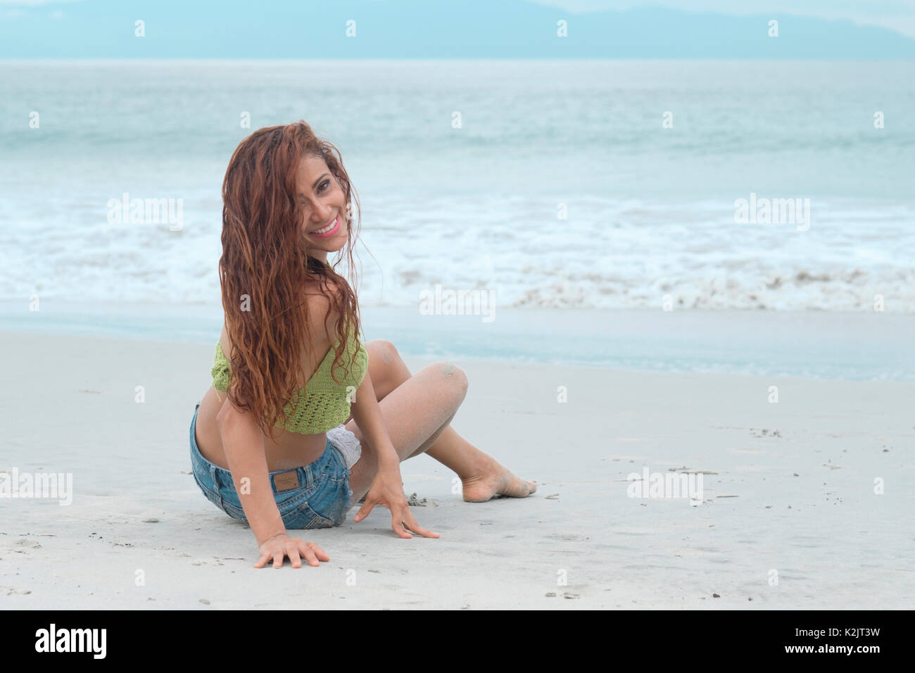 Barefoot woman sitting in sand at beach smiling while turning her body back Stock Photo