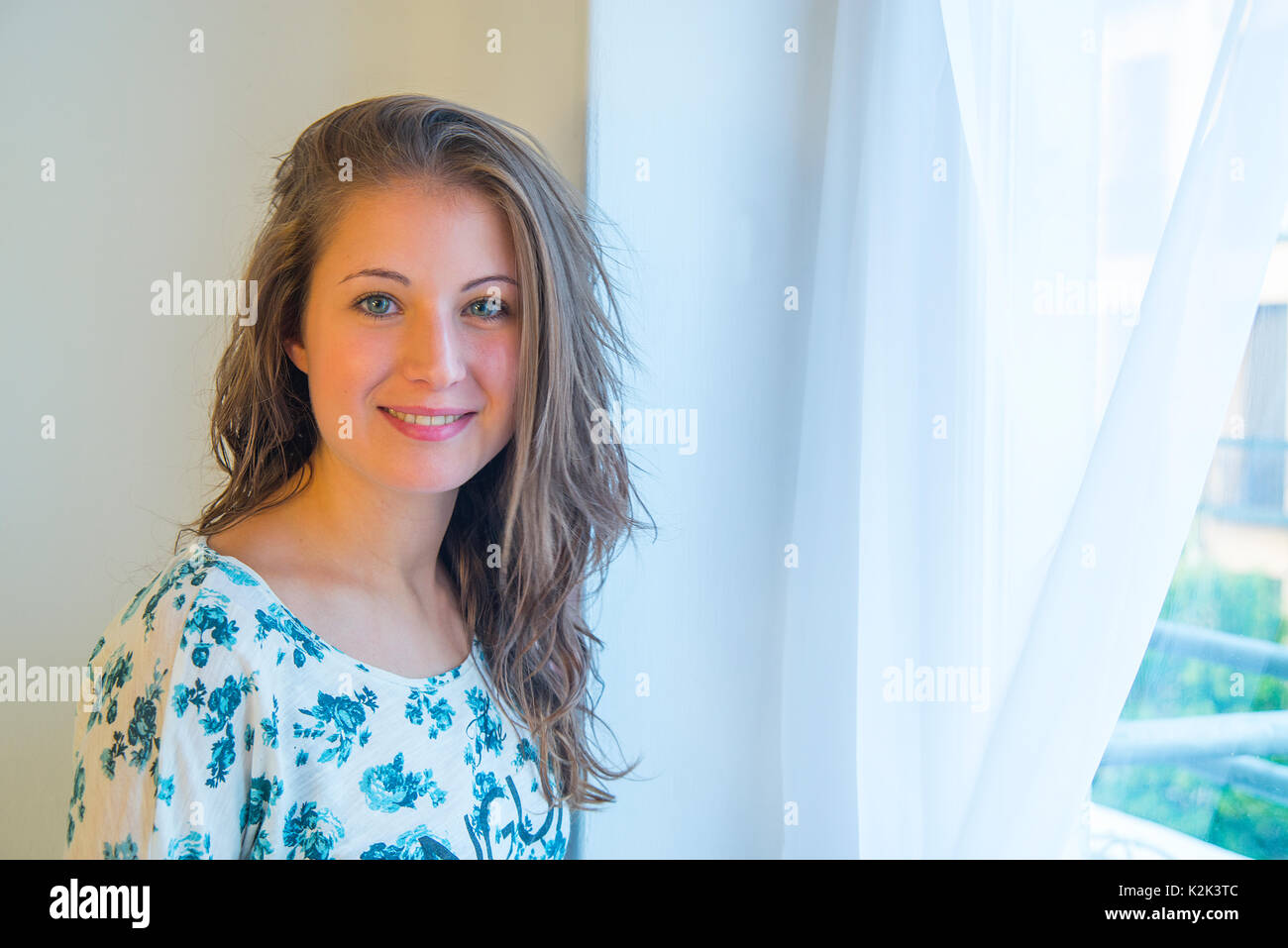 Young woman by the window, smiling and looking at the camera. Stock Photo