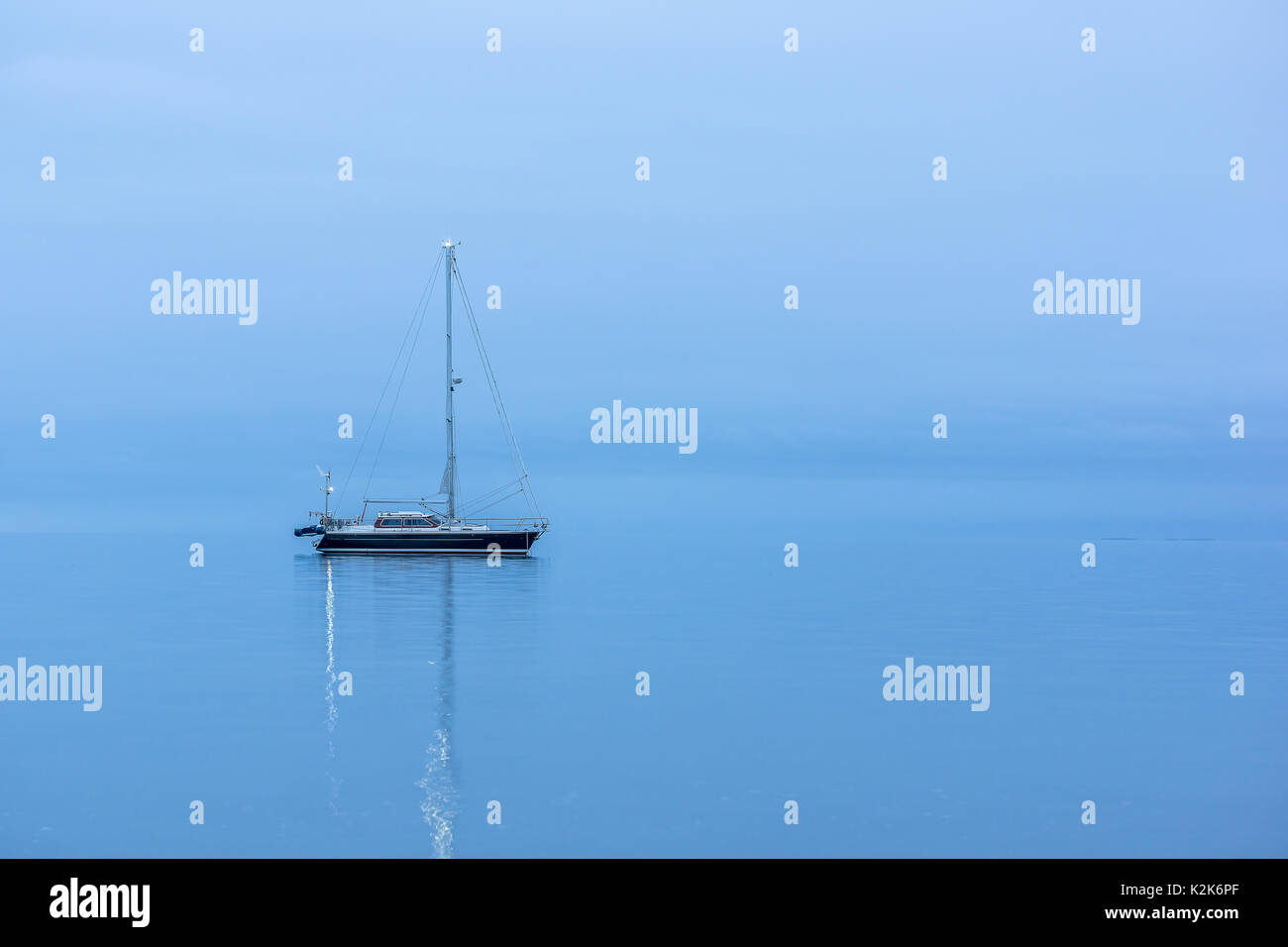 Lonely Black sailing boat in the ocean before sunrise, reflections in blue hour Stock Photo