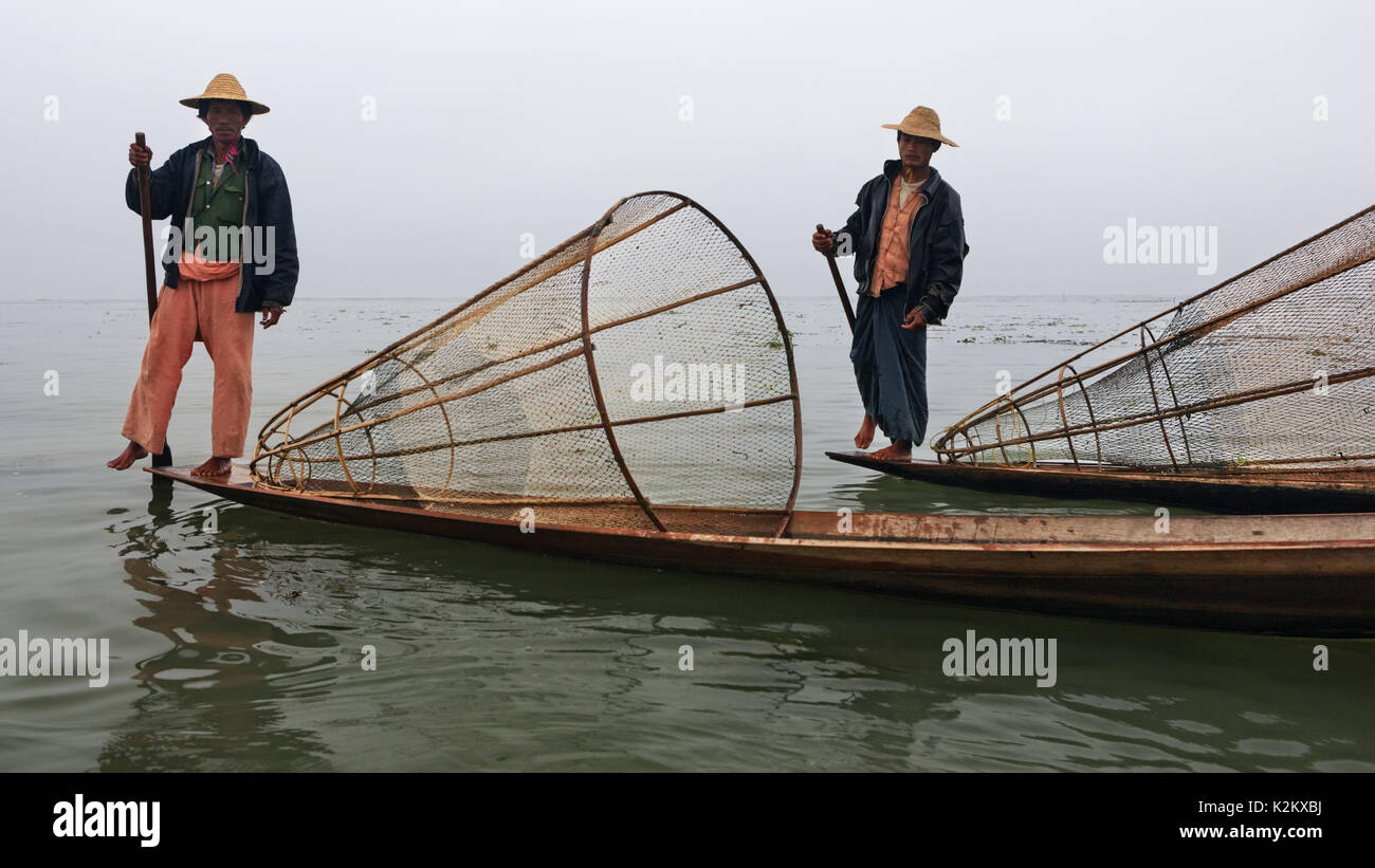 Inle Lake, Myanmar - January 04, 2007: Two local fishers with traditional Burmese fishing nets standing on stern of long boats and rowing oars by leg. Stock Photo