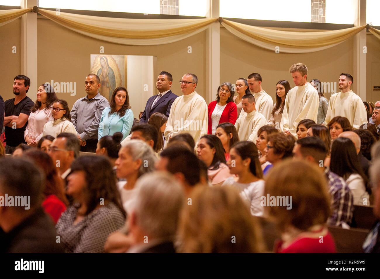 Multiracial baptism candidates and their sponsors line up beside the congregation at a baptism mass at an Orange, CA, Catholic cathedral. Note baptism robes and range of ages. Stock Photo