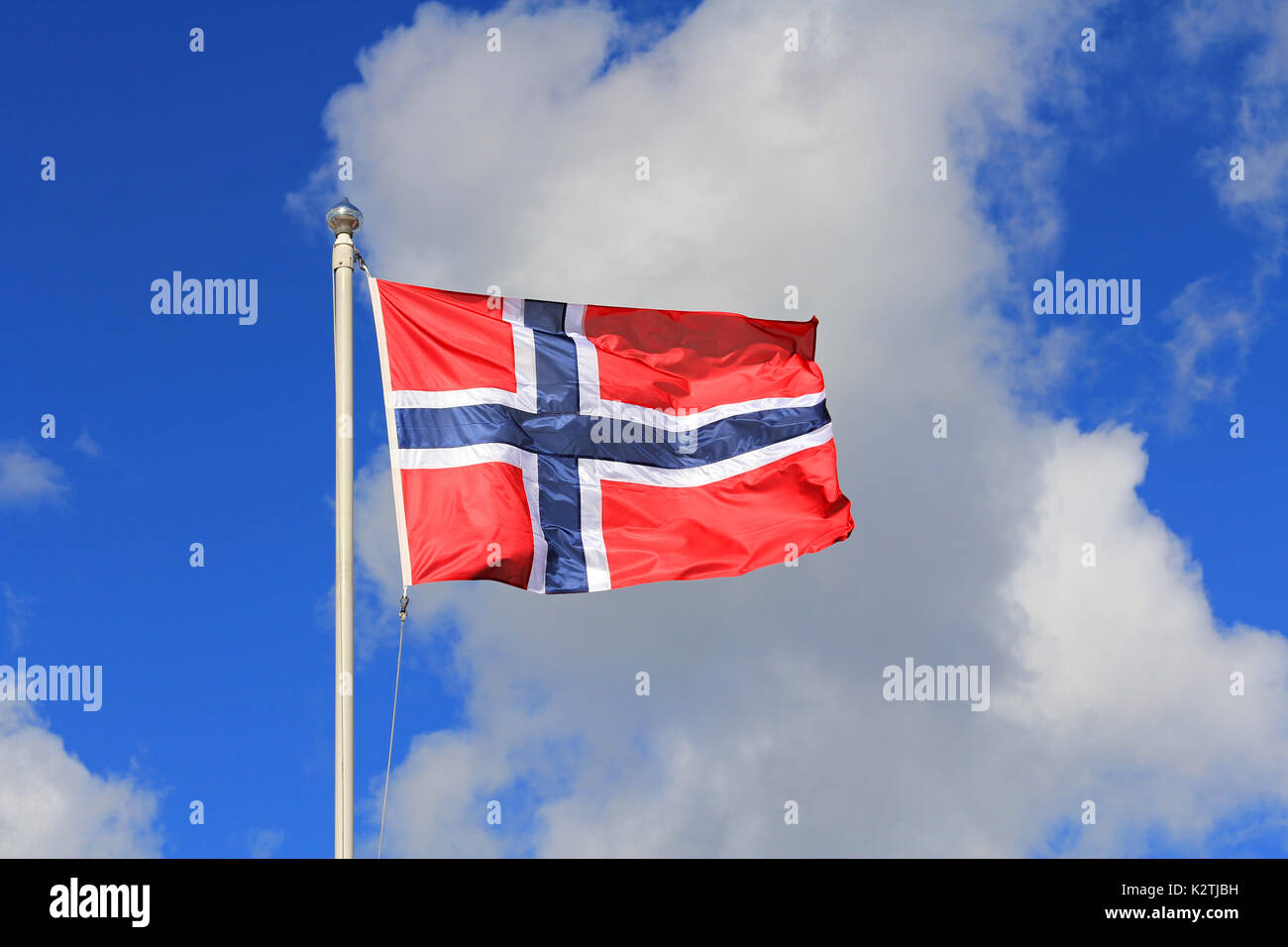 Flag of Norway against blue sky and white clouds. The flag of Norway is red with an indigo blue Scandinavian cross fimbriated in white. Stock Photo