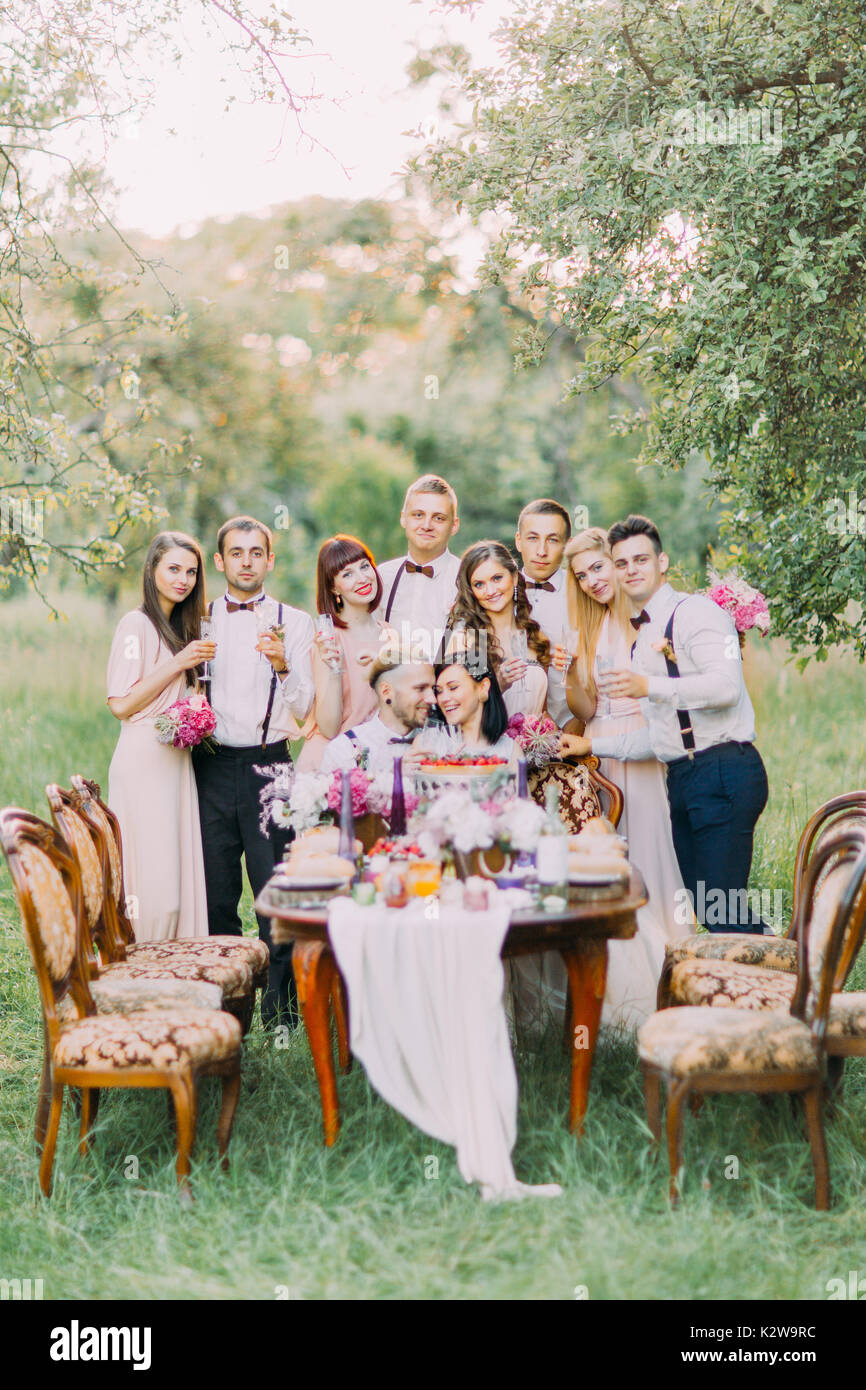 The vertical photo of the happy newlywed couple and the smiling guests standing behind them at the background of the spring green field. Stock Photo