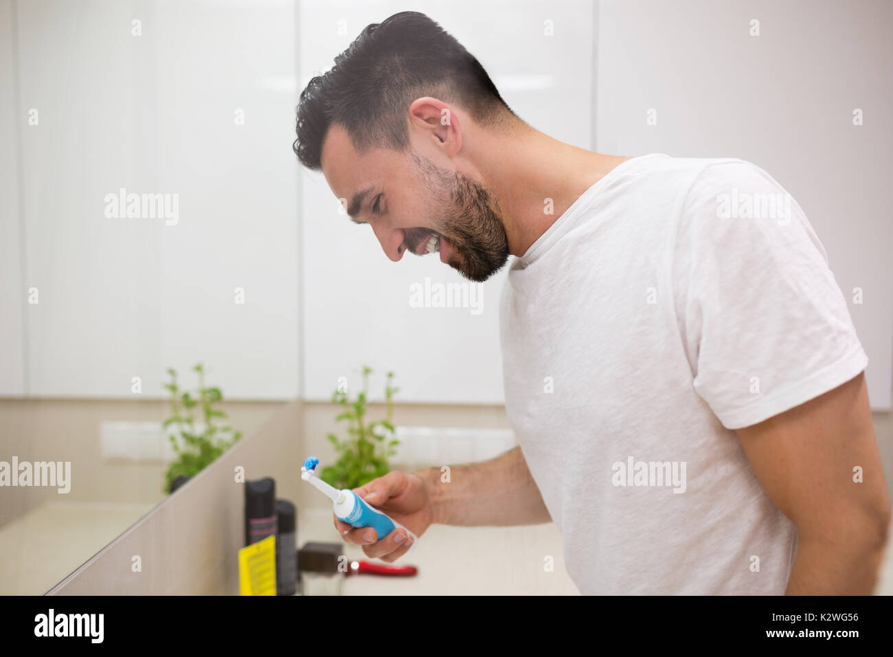 Joyful young man smiling during morning teeth brushing. He has black eyes and a beard. He is wearing white t-shirt. Stock Photo