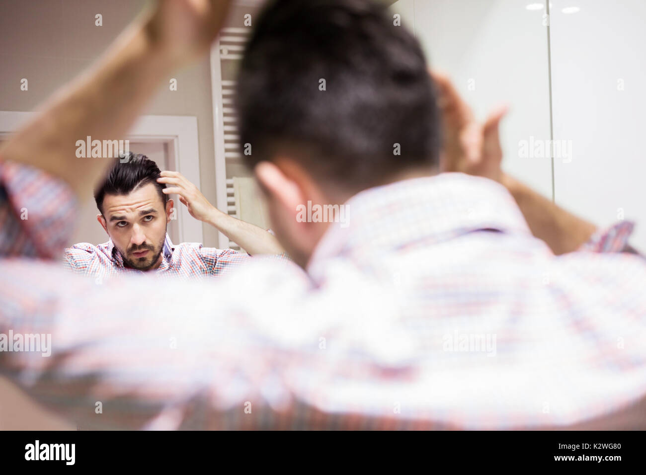 Young man applying hair cosmetics during hair caring. He is wearing shirt with checkered pattern. He is touching his black hair. Stock Photo