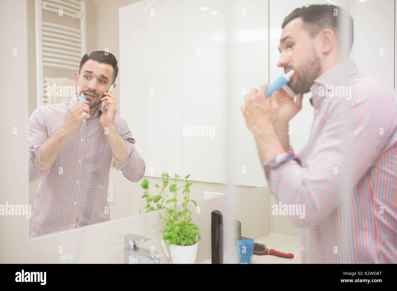 Man standing in bathroom, brushing teeth and having phone call. He is using electric toothbrush, looking away and wearing casual shirt with rolled up  Stock Photo