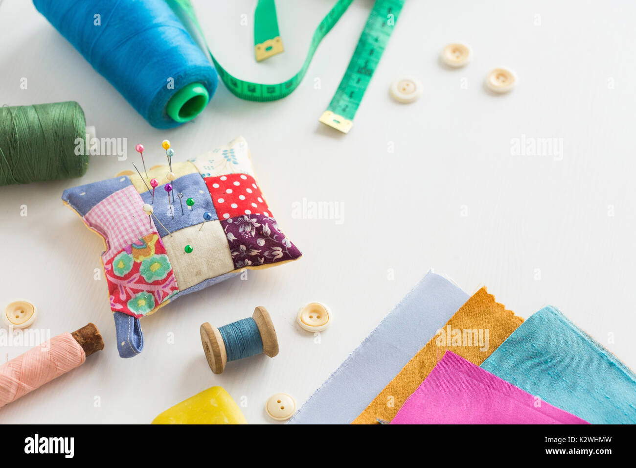 needlework, craft, sewing and tailoring concept - tools closeup on white desk, measuring meter, blue, pink and green threads in spools, scraps of colorful fabric, pincushion, white buttons and soap. Stock Photo