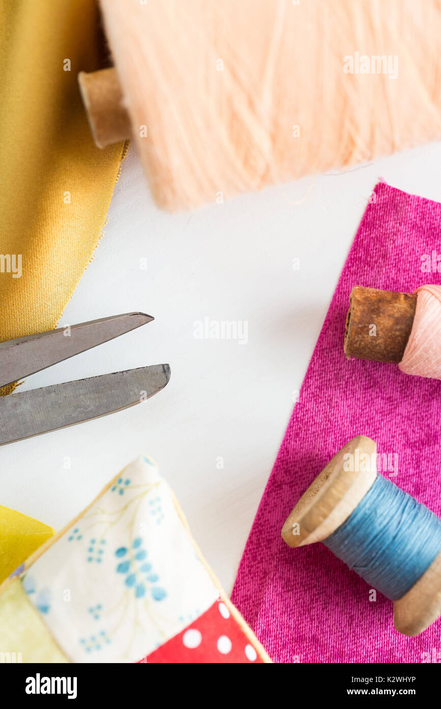 sewing, patchwork, tailoring and fashion concept - closeup tools on white work desk in studio, scissors, spools of blue and pink threads, pincushion, pieces of colored patchwork fabric, vertical. Stock Photo
