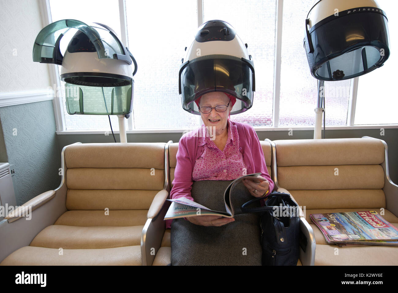 Elderly pensioner having her hair dried in a hood dryer wearing curlers at a ladies hairdresser's salon, England, United Kingdom Stock Photo