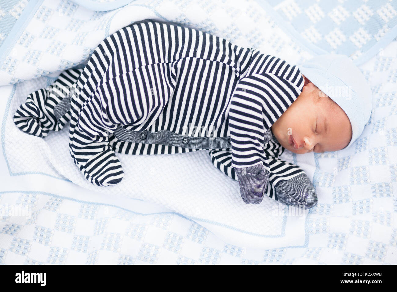 Sleeping newborn baby in striped romper, mittens and beanie lies on side in blue coverlet. Stock Photo