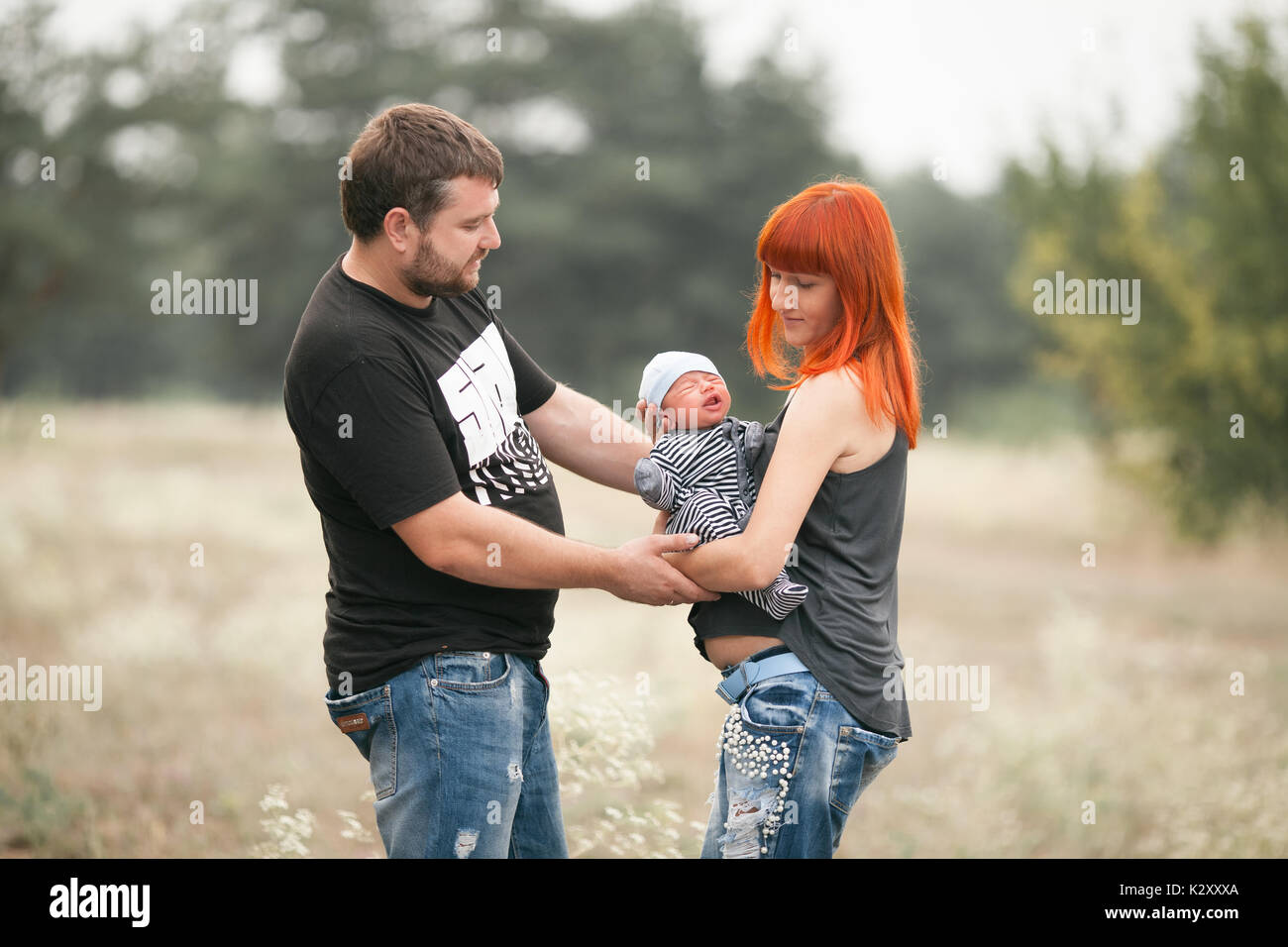 Happy young family with newborn baby on walk in park. Mother holds in her hands crying newborn baby, father stands near. Stock Photo