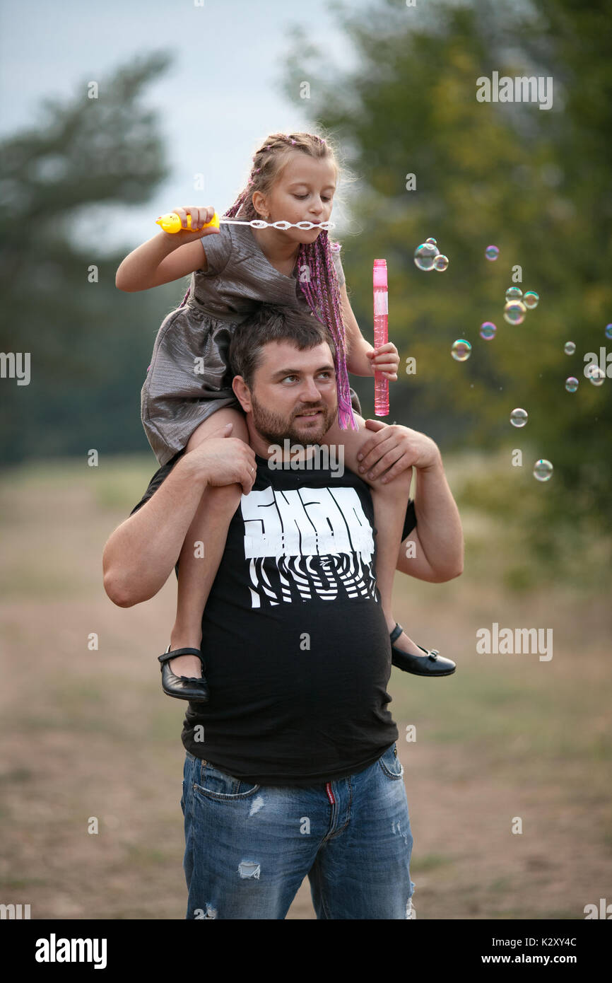 Happy father with daughter blowing soap bubbles on walk in park. Dad put child girl on his shoulders. Stock Photo