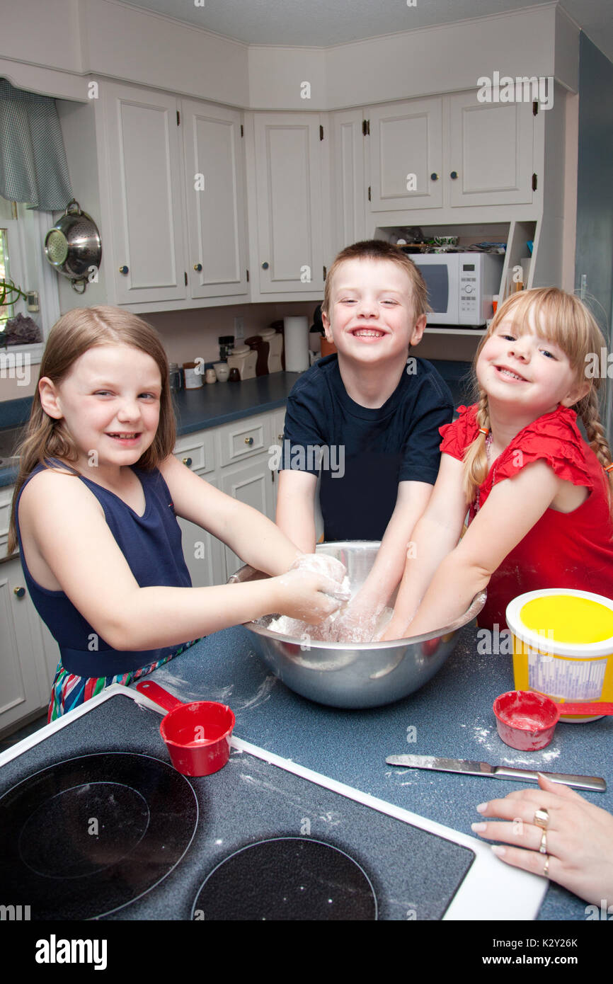 Three happy kids in kitchen with their hands in a big bowl full of dough at home Stock Photo