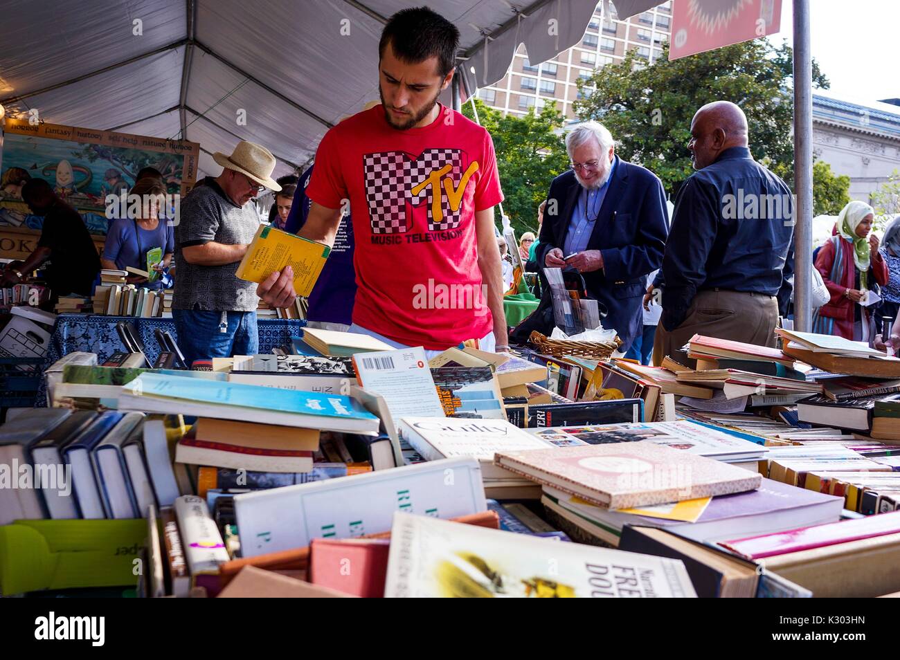 A young man wearing a MTV t-shirt exploring a large pile of books among other people exploring books at a tent at the Baltimore Book Festival, Baltimore, Maryland, September, 2013. Stock Photo