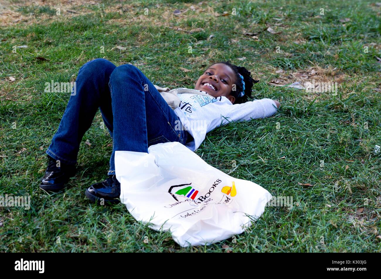 A young girl smiling and laying down in the grass with a plastic bag at the Baltimore Book Festival in Baltimore, Maryland, September, 2013. Stock Photo
