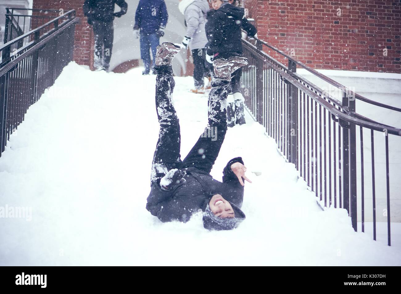 A student sits in the snow with legs in the air after sliding down the snowy staircase, while other students dressed in snow gear stand at the top of the staircase, on a snow day at Johns Hopkins University, Baltimore, Maryland, 2016. Stock Photo