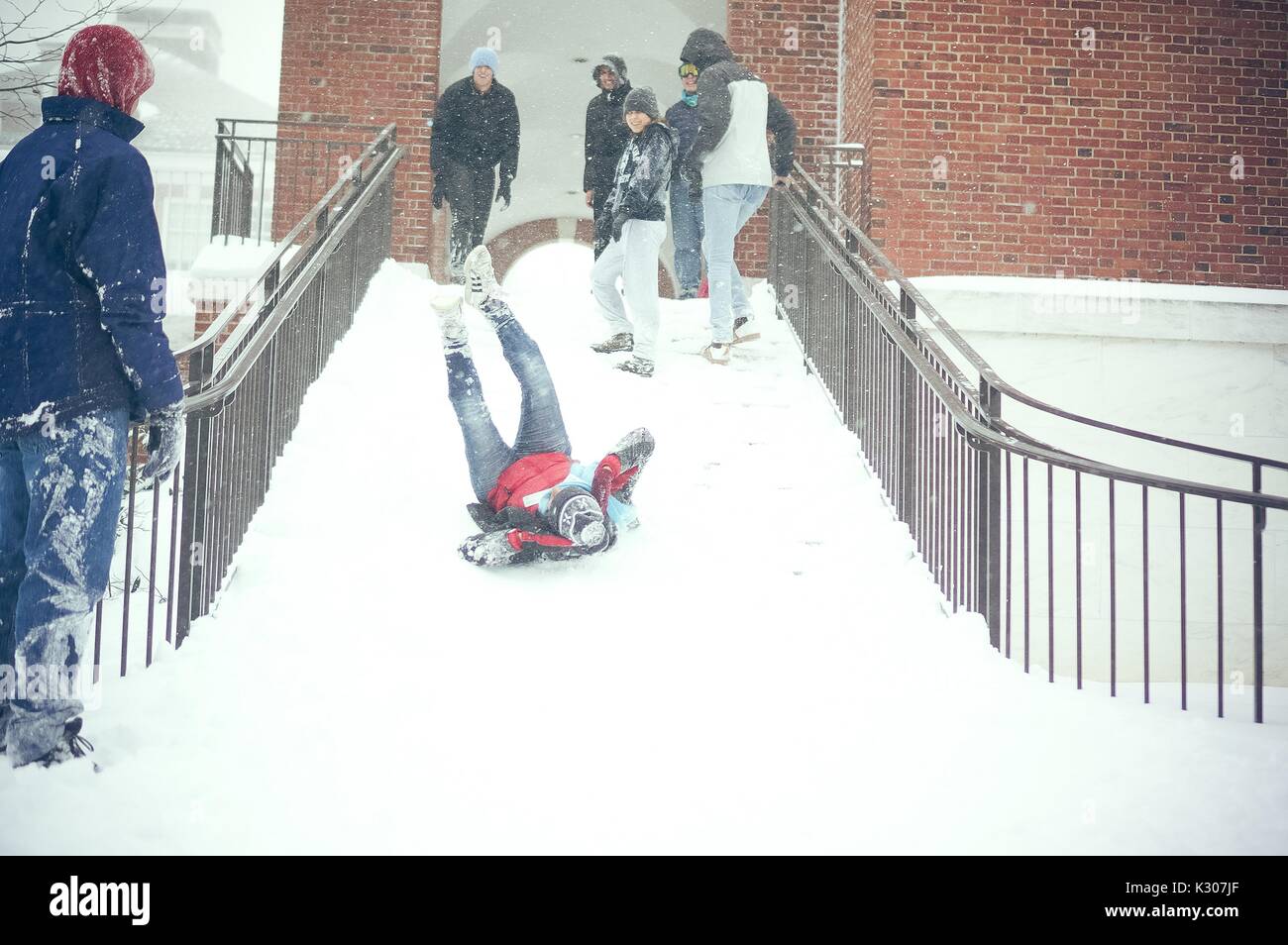 A student slides down the snowy stairs with legs in air and arms around head, while students dressed in snow gear laugh and cheer him on from the top of the staircase, during a snow day at Johns Hopkins University, Baltimore, Maryland, 2016. Stock Photo