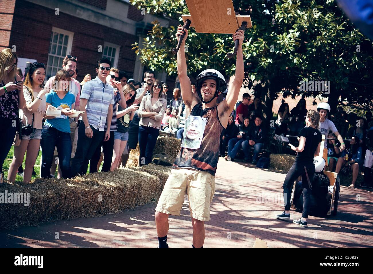 A crowd watches from the sidelines as a male student wearing tank top and helmet smiles with his arms in the air, wielding a wooden contraption, signaling victory during a carnival game at Spring Fair, a student-run spring carnival at Johns Hopkins University, Baltimore, Maryland, April, 2016. Courtesy Eric Chen. Stock Photo