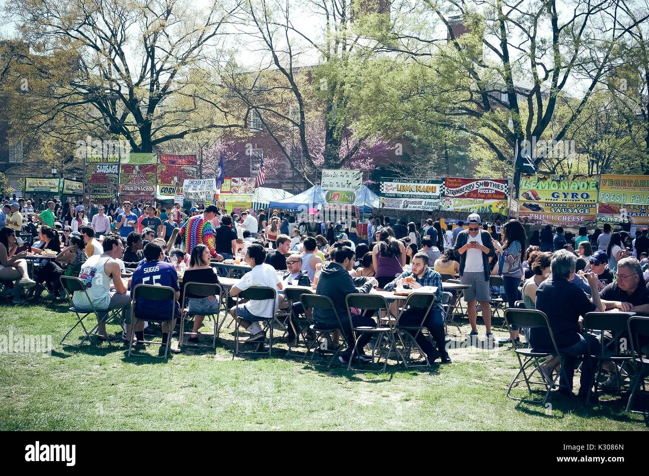 Nearly a hundred people sit eating outdoors on the grass at tables or stand in line for dozens of food trucks during Spring Fair, a student-run spring carnival at Johns Hopkins University, Baltimore, Maryland, April, 2016. Courtesy Eric Chen. Stock Photo