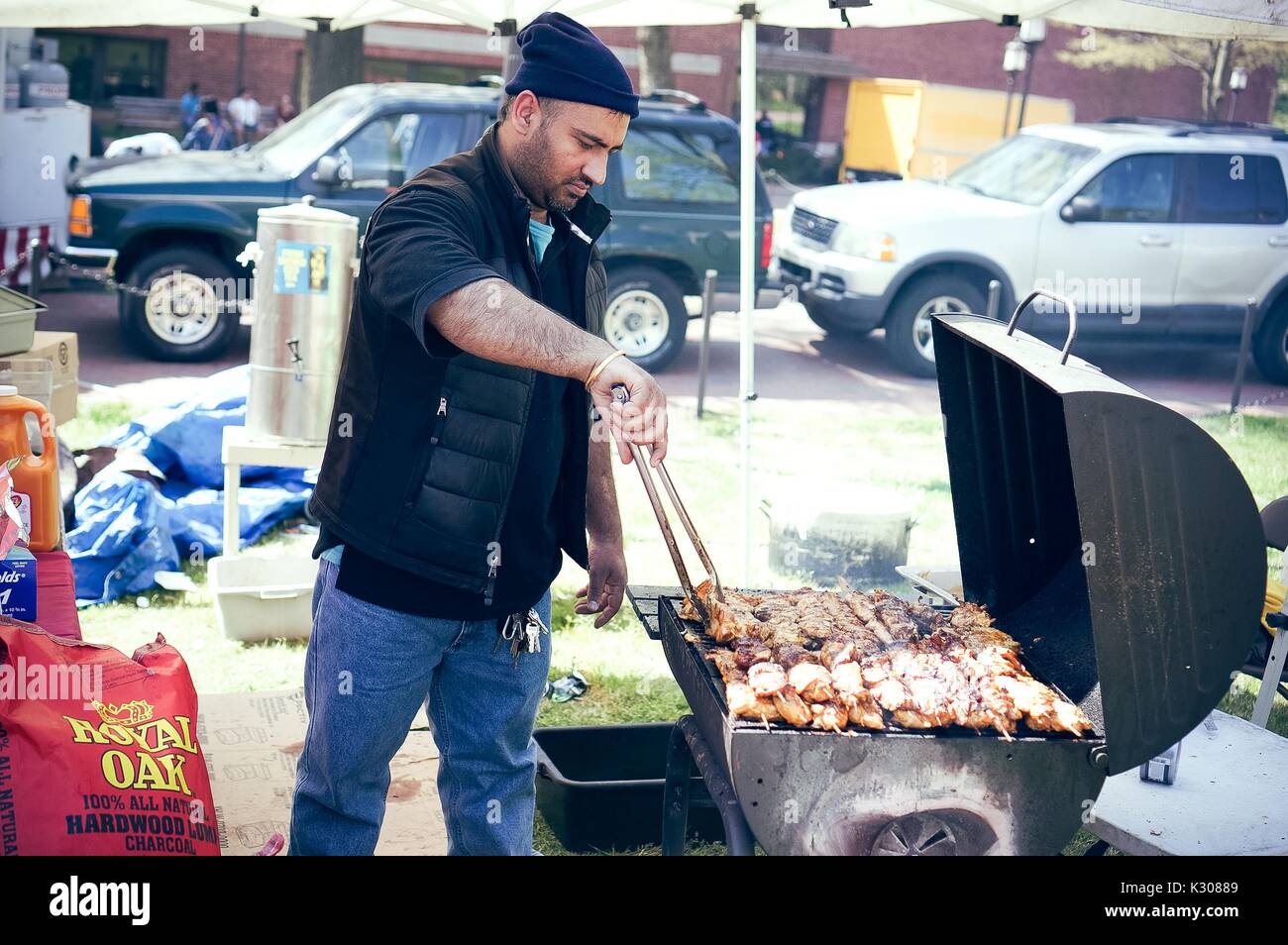 A man wearing beanie and vest stands in front of a grill, cooking chicken on skewers using large tongs, at a food booth during Spring Fair, a student-run spring carnival at Johns Hopkins University, Baltimore, Maryland, April, 2016. Courtesy Eric Chen. Stock Photo