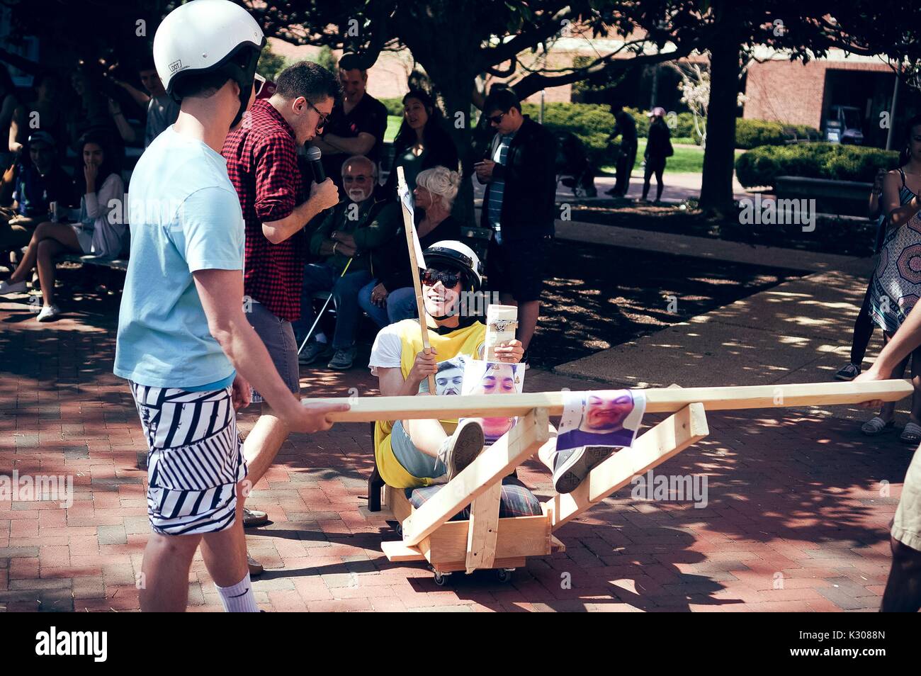 Two male students wearing helmets carry a third student inside a wooden contraption during a carnival game, while a male student narrates with a microphone and onlookers watch from the shade, at Spring Fair, a student-run spring carnival at Johns Hopkins University, Baltimore, Maryland, April, 2016. Courtesy Eric Chen. Stock Photo