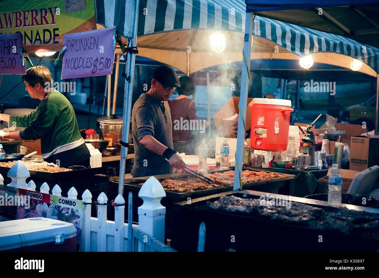 Two men stand in front of large grills cooking chicken on skewers at a food booth at dusk, during Spring Fair, a student-run spring carnival at Johns Hopkins University, Baltimore, Maryland, April, 2016. Courtesy Eric Chen. Stock Photo
