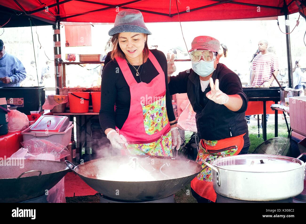 A woman wearing hat and apron stirs noodles in a giant wok, while a man beside her throws up peace signs with gloved hands, behind a food booth at Spring Fair, a student-run spring carnival at Johns Hopkins University, Baltimore, Maryland, April, 2016. Courtesy Eric Chen. Stock Photo