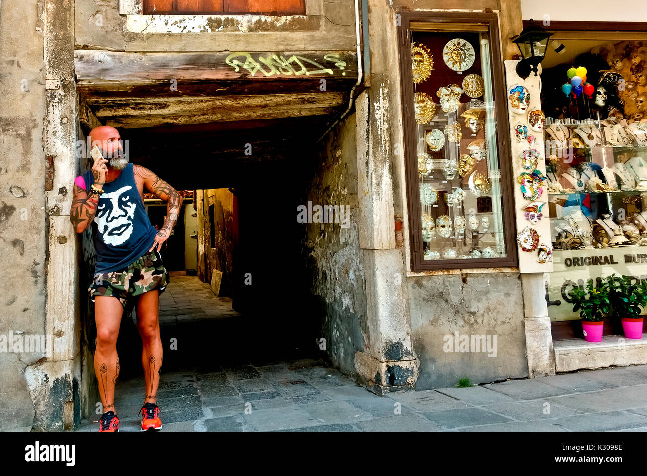 Tatooed, bearded, bald headed man wearing tank top and military short pants, leaning against wall, talking on mobile phone. Venice, Italy, Europe, EU. Stock Photo