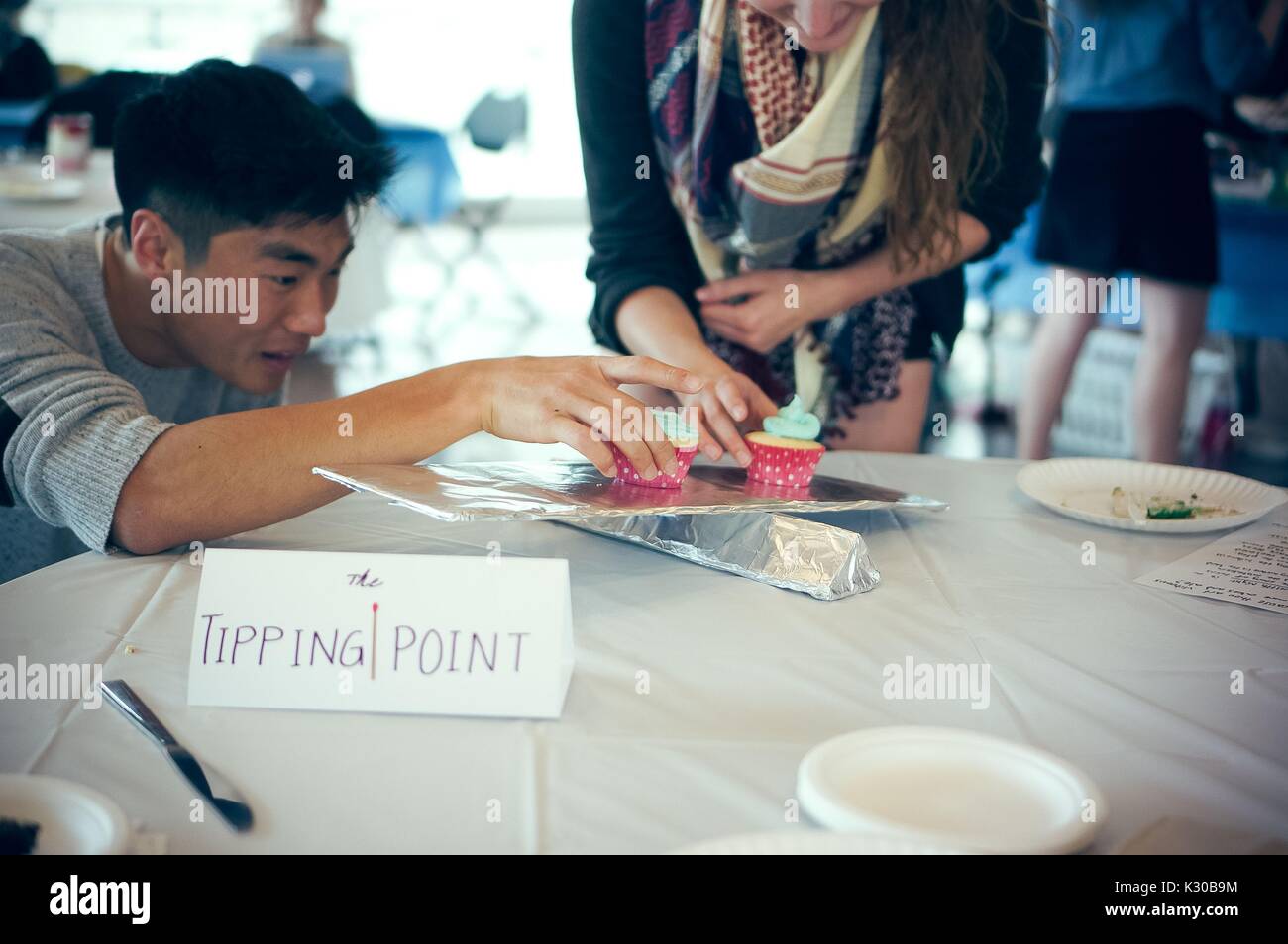 Two undergraduate students looking at an aluminum scale with cupcakes on it representing 'The Tipping Point' by Malcolm Gladwell at the Edible Book Festival at Johns Hopkins University, Baltimore, Maryland, March 31, 2016. Courtesy Eric Chen. Stock Photo