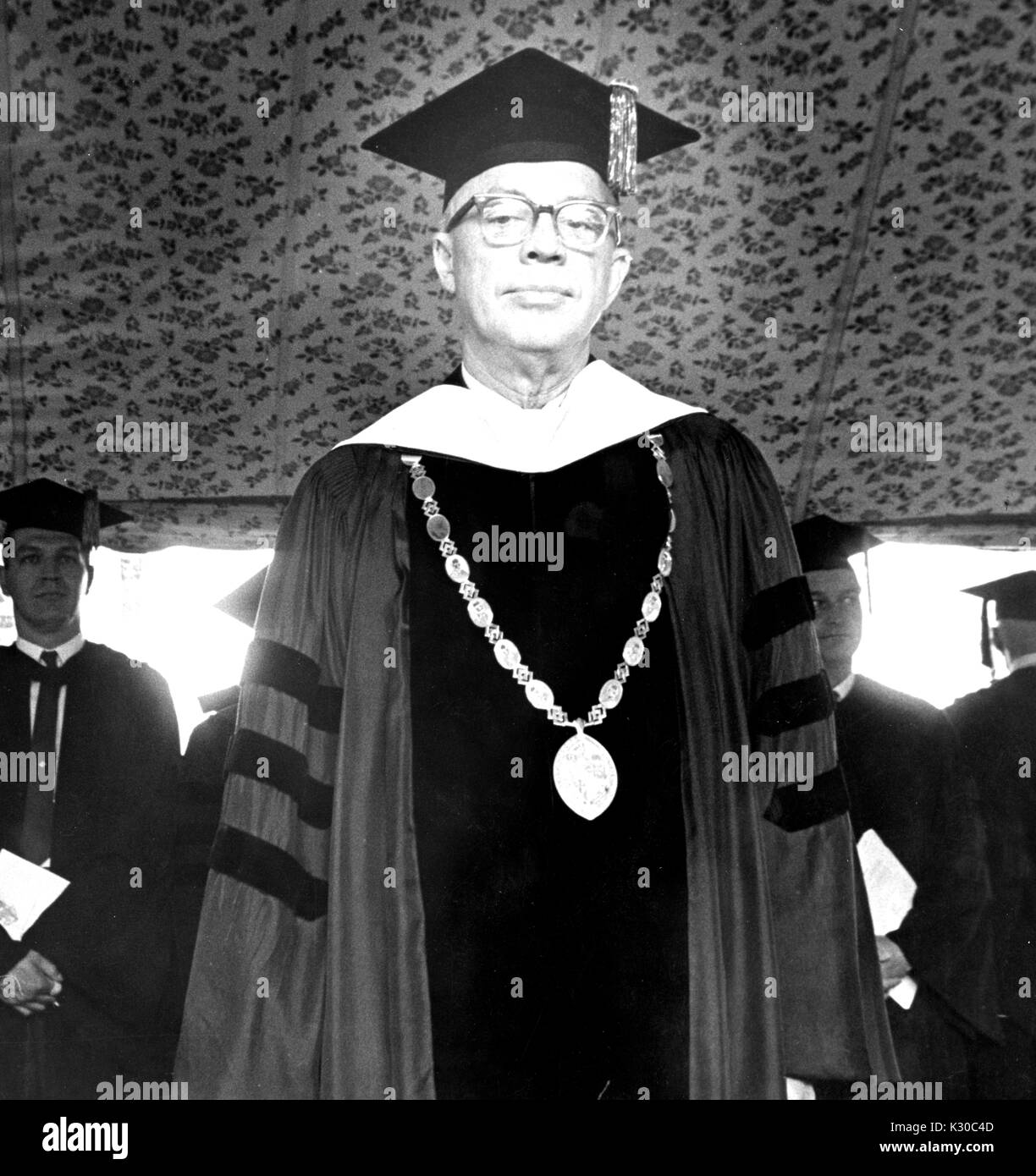 On Commencement Day, President of Johns Hopkins University Milton Stover Eisenhower stands on stage wearing graduation garb and medal, looking solemn, with graduates standing behind him with hands together, Baltimore, Maryland, June 13, 1967. Stock Photo