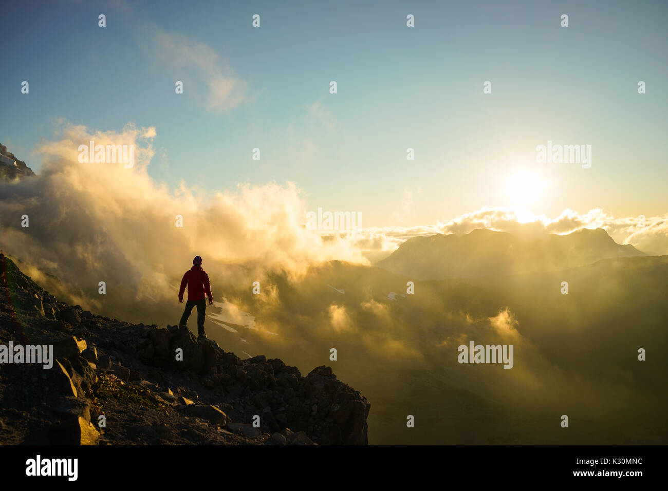 A hiker is standing on the mountain with clouds during sunset/sunrise at Mount Rainier National Park, Washington. Stock Photo