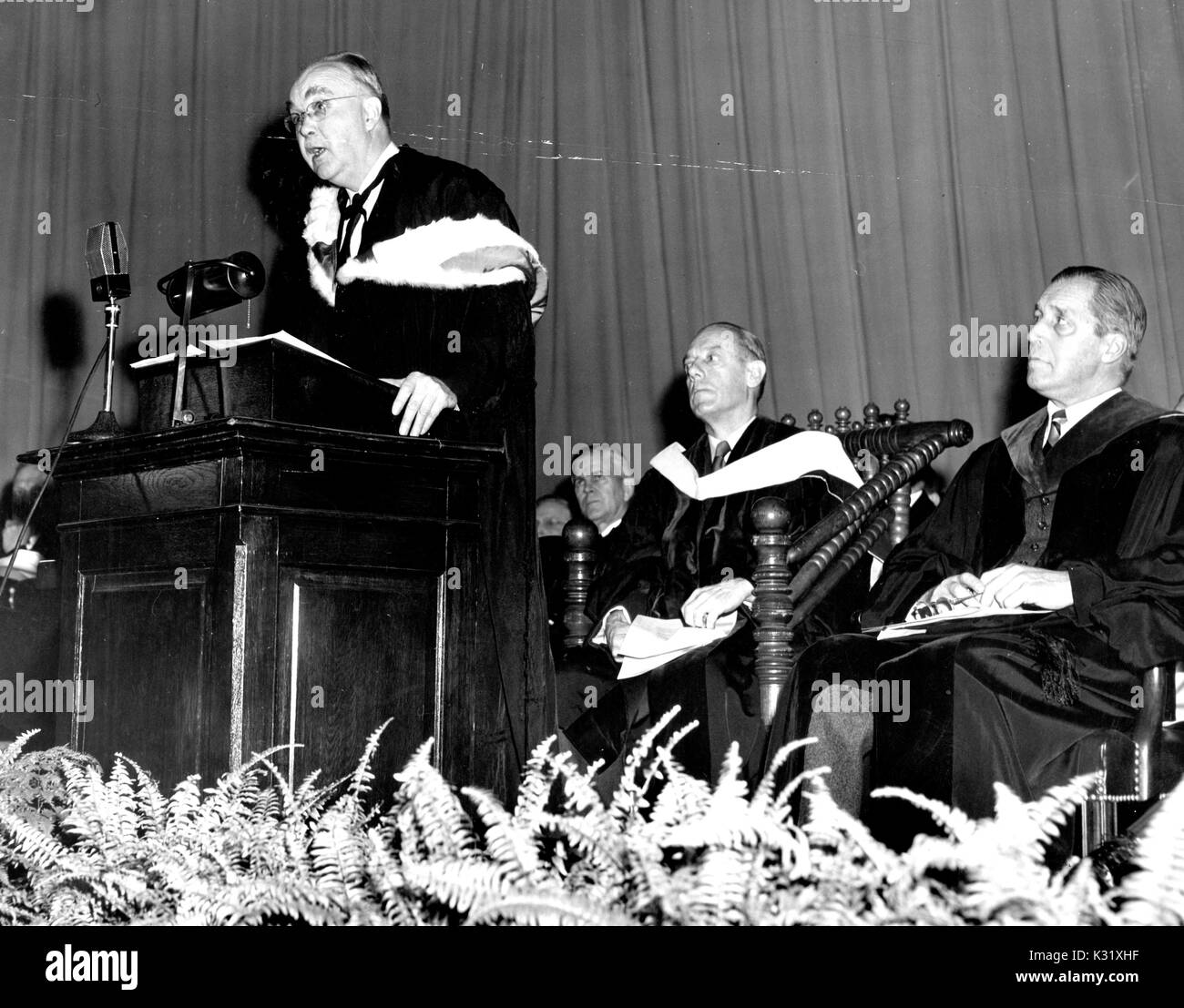 Commemoration Day 1951 at Johns Hopkins University, with humanities scholar Henry Allen Moe standing at the podium to the left giving a speech to graduating students, while biophysist Detlev Wulf Bronk and chaplain Thomas Guthrie Speers seated behind him on the stage, all wearing graduation garb, Baltimore, Maryland, February 22, 1951. Stock Photo