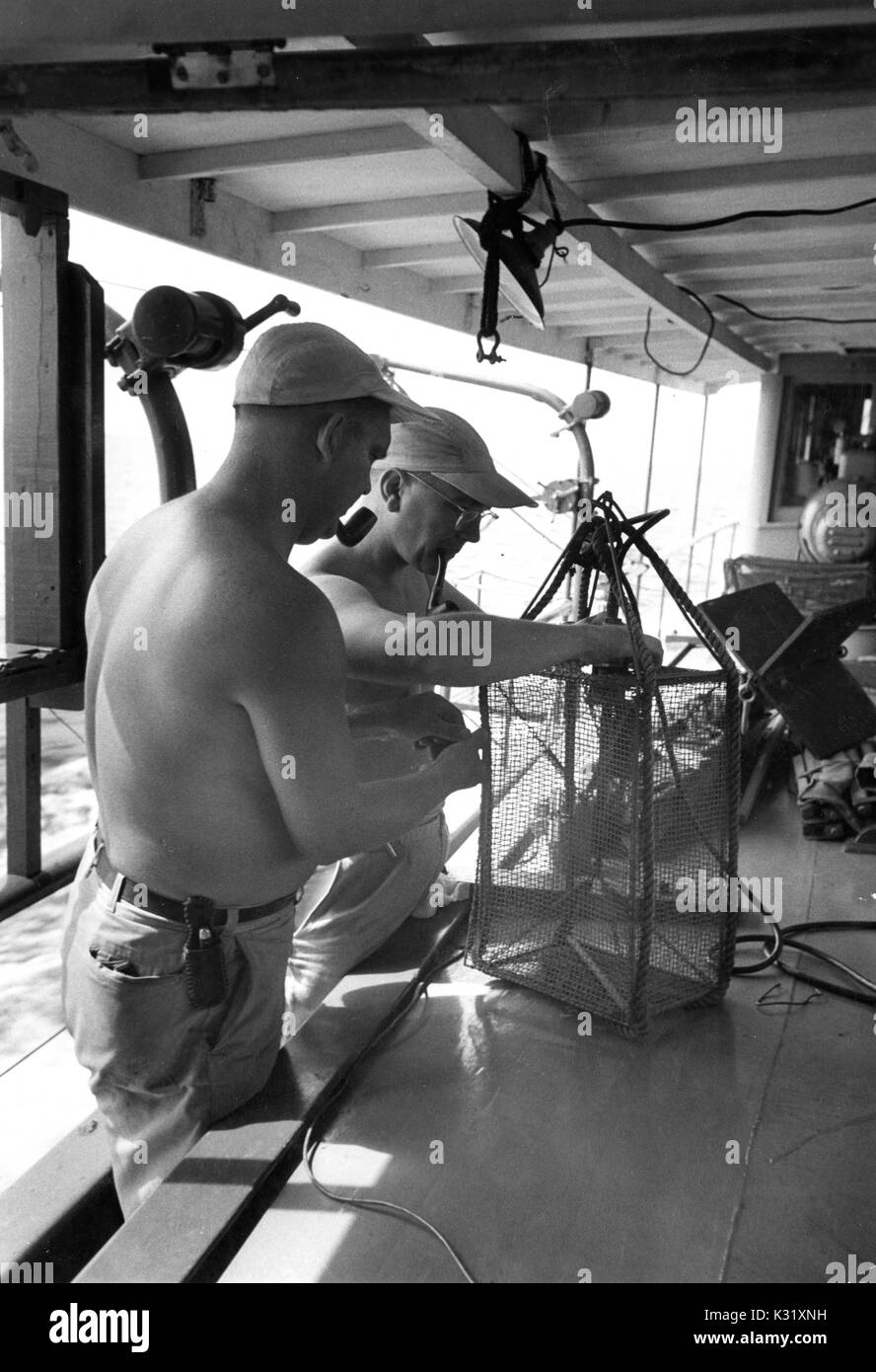 Two shirtless research scientists with tobacco pipes in mouth, adjusting a  device to test the water's conductivity and temperature, aboard the Joanbar  II, a research vessel working for the Chesapeake Bay Institute