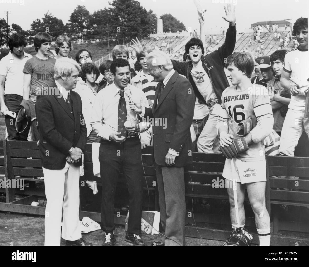 A man is speaking into a microphone during an interview for the 8th Annual NCAA Division One Championships, while spectators watch and try to make a scene for the camera, with a Johns Hopkins lacrosse player standing by, 1978. Stock Photo