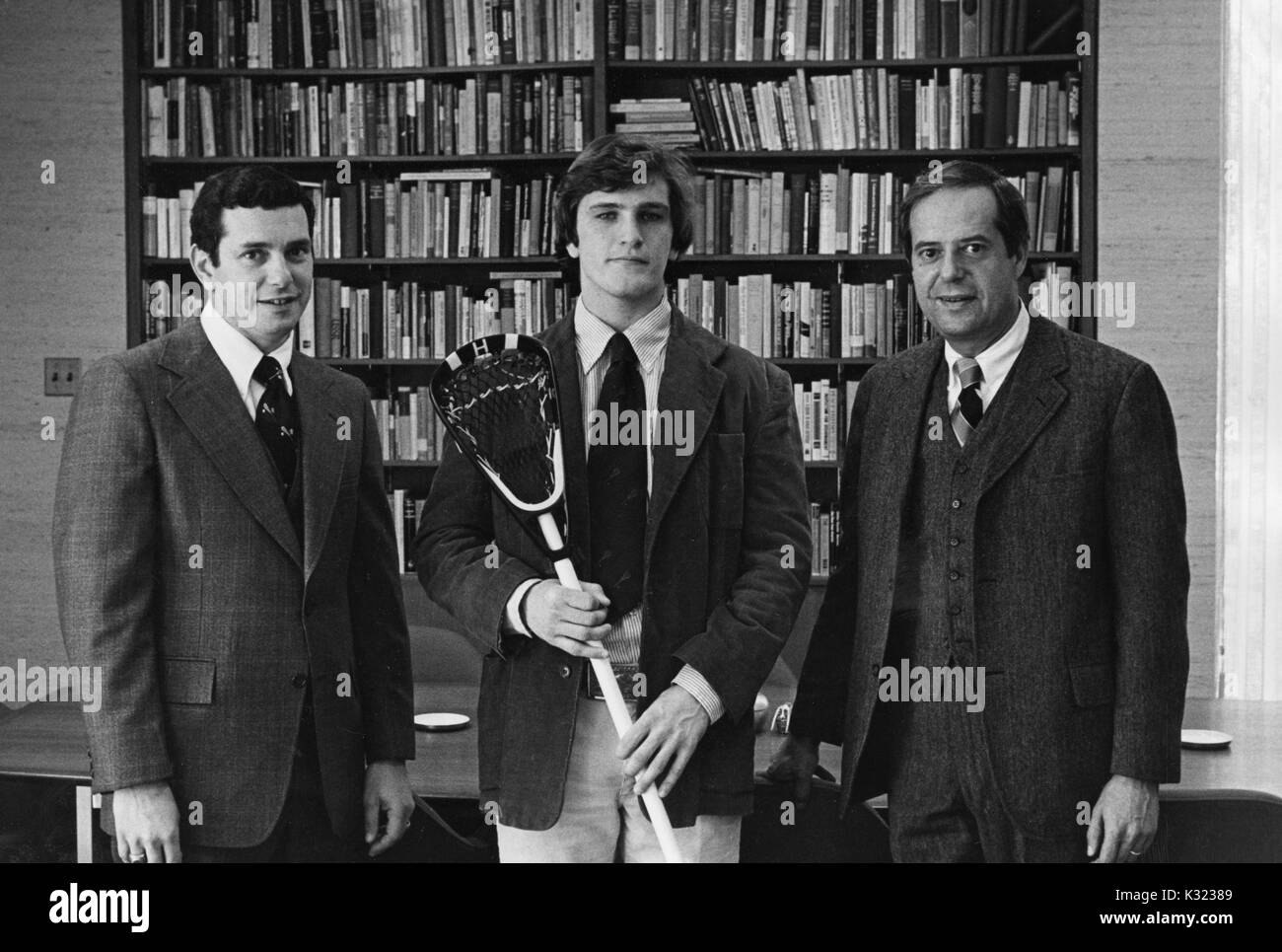 Three time All-American lacrosse player for Johns Hopkins University, Mike O'Neill, wears a blazer and holds his stick to pose with head coach for men's lacrosse, Henry Ciccarone, to his left, and Steven Muller, president of Johns Hopkins University, to his right, in front of a wall of books, Baltimore, Maryland, May, 1978. Stock Photo