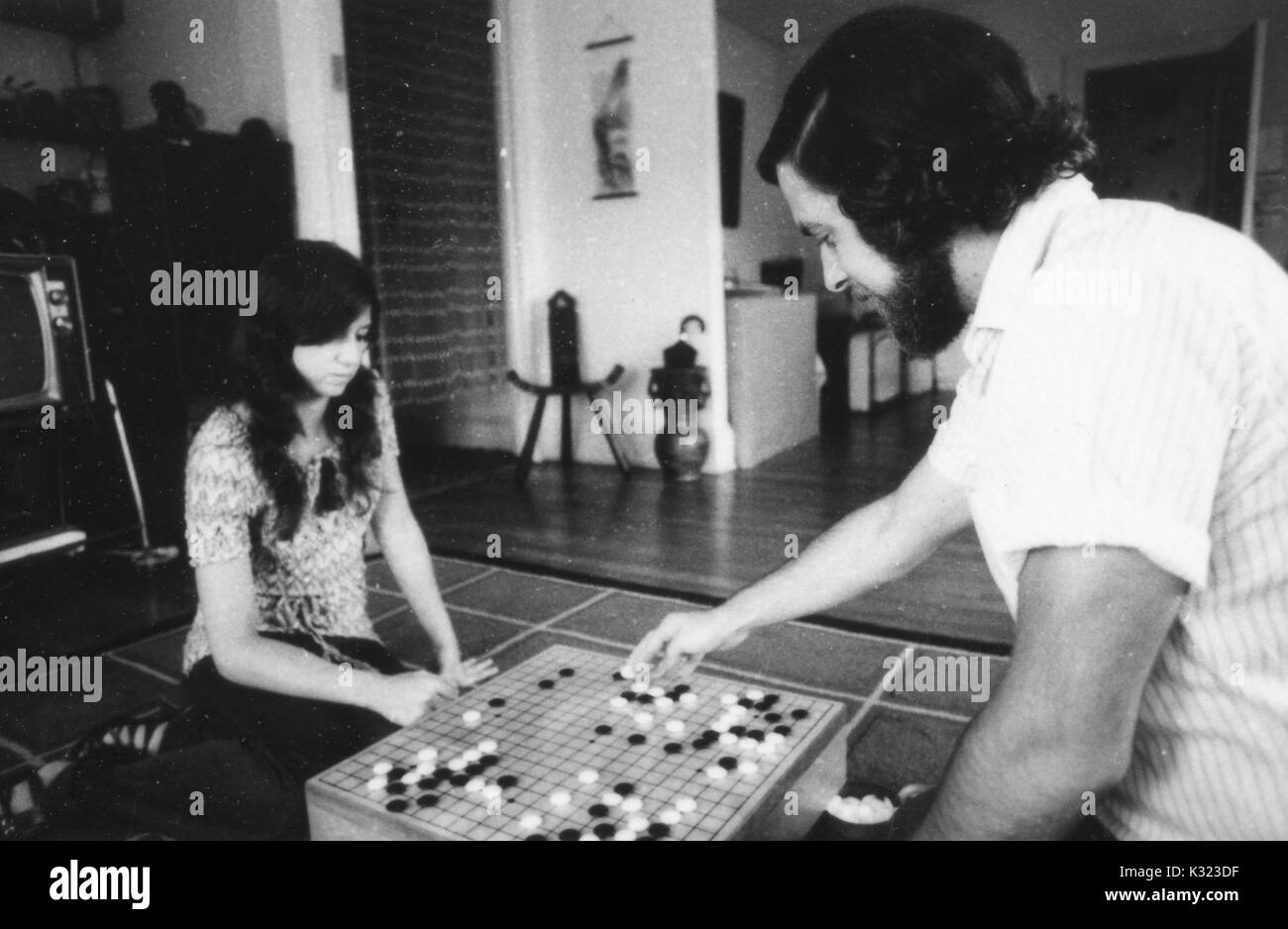 A male and female student sit on opposite sides of a grid board engaged in a game of Go, an old Asian strategy game, with white and black stones arranged on the grid, 1970. Stock Photo