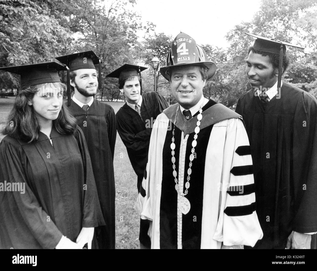 After the commencement ceremony, four graduates -- including politician and MSNBC analyst Michael Steele and Arts and Science graduate Robert Friedman -- wearing caps and gowns pose outdoors with President Steven Muller -- German-American political science scholar and President of Johns Hopkins University (1972-1990) -- wearing an academic robe, the University medal, and a fireman's helmet that reads 'Go-For 4 Jays, ' Baltimore, Maryland, June, 1981. Stock Photo