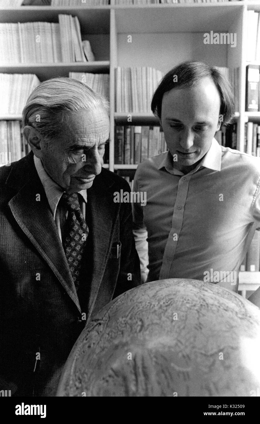 In a building for Earth and Planetary Science at Johns Hopkins University, famed geophysicist Walter M Elsasser wears a suit and stands beside department professor Peter L Olsen as they examine a large globe together in front of a bookcase filled with research and textbooks, Baltimore, Maryland, 1975. Stock Photo