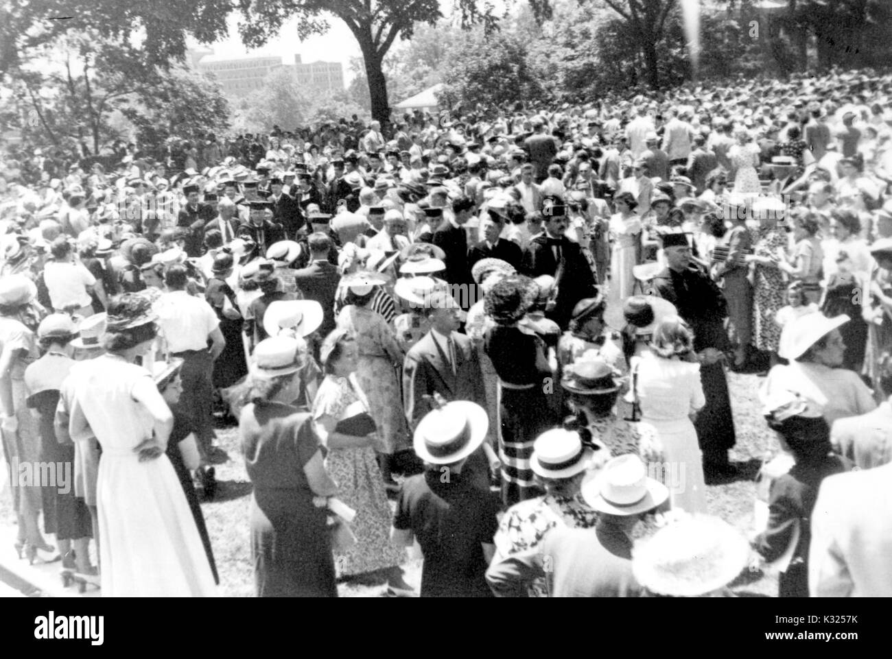 On Commencement Day, a large crowd of family, friends, and guests stand outdoors in clusters watching an aisle of graduates and faculty members proceed together wearing caps, gowns, and robes, Johns Hopkins University, Baltimore, Maryland, June, 1949. Stock Photo
