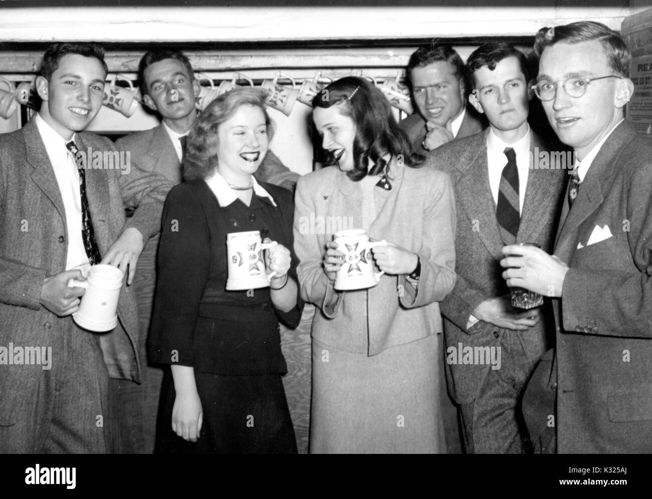 At a party hosted by the Delta Phi fraternity at Johns Hopkins University, brothers stand in a group wearing suits next to their girlfriends, everyone smiling and drinking beer from tall mugs, in a fraternity house basement right off campus, Baltimore, Maryland, March, 1947. Stock Photo