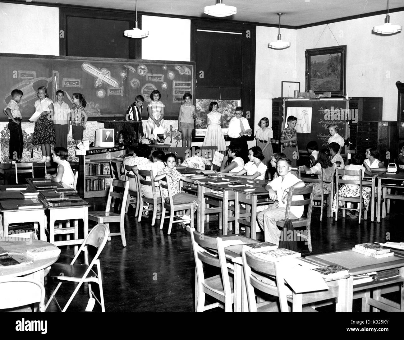 At an elementary demonstration school of Johns Hopkins University, students in Mr Fuller Strawbridge's sixth-grade Space Academy sit at desks while a line of students presents their work at the front of the classroom, with a blackboard filled with space illustrations behind them, and Mr Strawbridge at the end of the line directing the activities, Baltimore, Maryland, June, 1955. Stock Photo