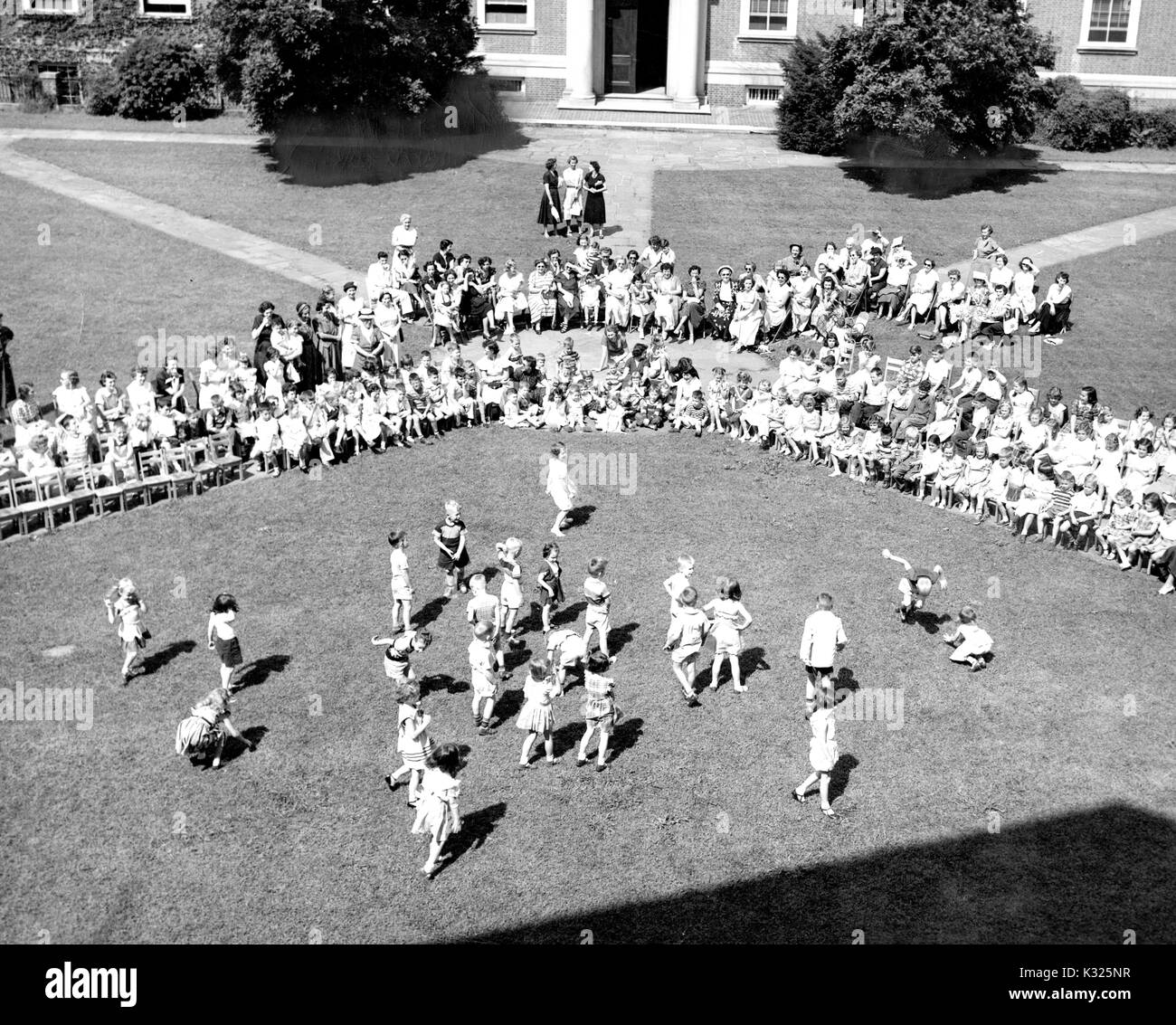 At the end of the school year for a demonstration school at Johns Hopkins University, young boys and girls put on a show in the grass on a sunny day, happily skipping and dancing in front of an audience made up of classmates, teachers, and parents sitting and standing outside of an ivy-covered campus building on an open quadrangle, Baltimore, Maryland, July, 1950. Stock Photo
