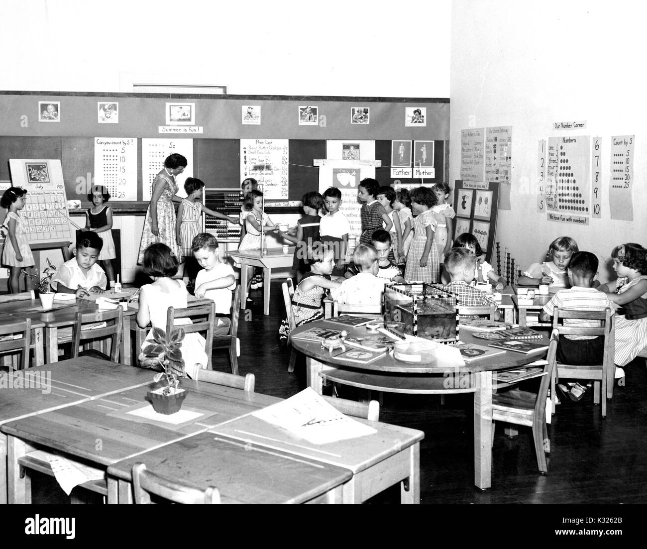 At an elementary demonstration school of Johns Hopkins University, students in Ms Ethel Brand Ford's first grade class sit at tables performing school work with crayons and workbooks, while two girls give a lesson with a calendar at the front of the room, beside Ms Ford who helps a line of students waiting to use the freestanding abacus for mathematics, Baltimore, Maryland, June, 1955. Stock Photo
