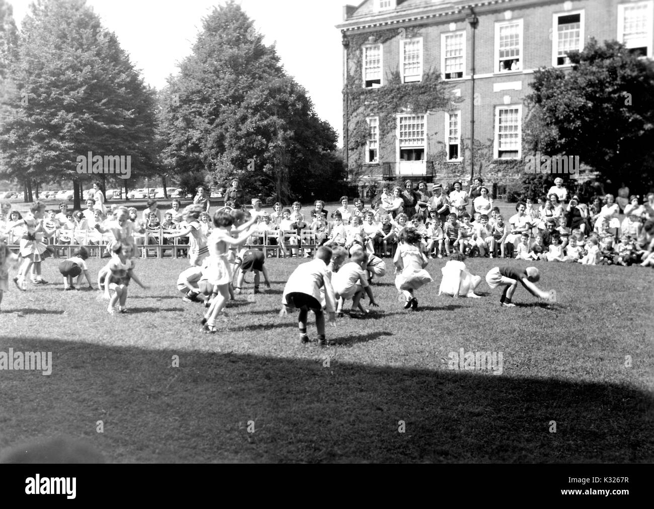 At the end of the school year for a demonstration school at Johns Hopkins University, young boys and girls put on a show in the grass on a sunny day, dancing and squatting down in front of an audience made up of classmates, teachers, and parents standing and sitting outside of an ivy-covered campus building, Baltimore, Maryland, July, 1950. Stock Photo