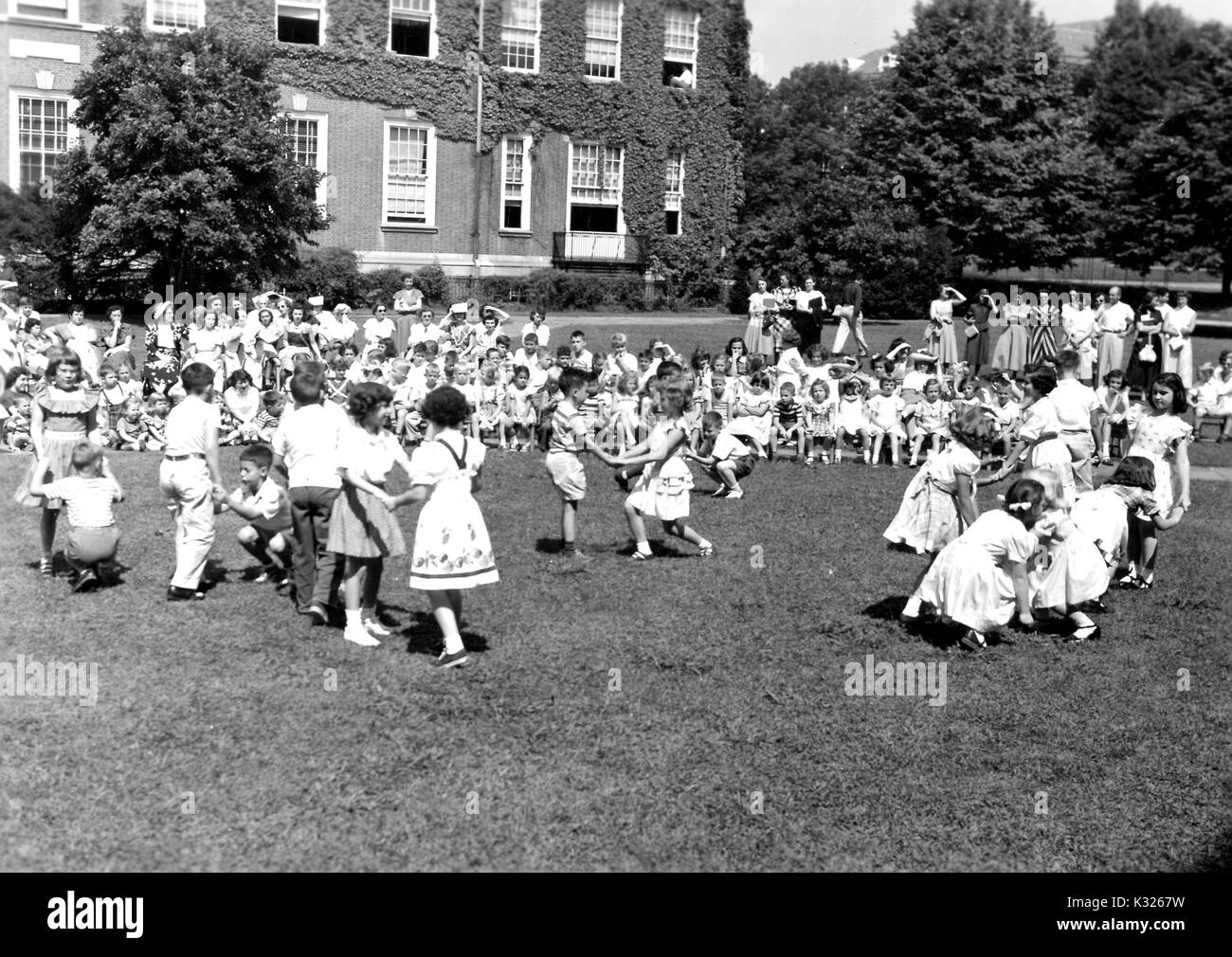 At the end of the school year for a demonstration school at Johns Hopkins University, young boys and girls put on a show in the grass on a sunny day, happily skipping and holding hands in front of an audience made up of classmates, teachers, and parents sitting and standing outside of an ivy-covered campus building, Baltimore, Maryland, June, 1950. Stock Photo