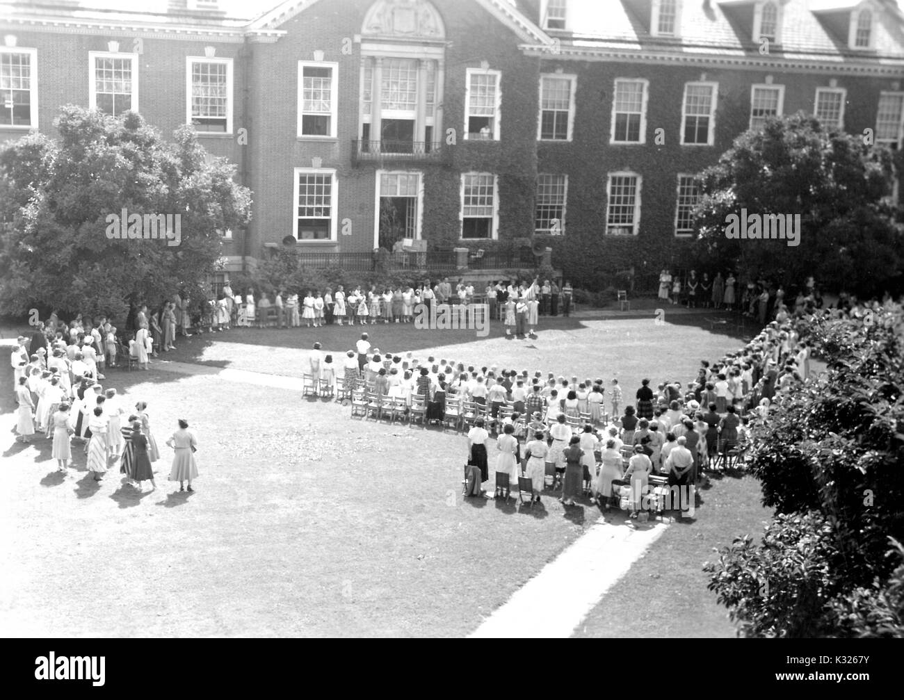 At the end of the school year for a demonstration school at Johns Hopkins University, parents and teachers stand in front of rows of chairs outside of an ivy-covered campus building, waiting for the young boys and girls to begin the show they will perform to close out the year, in the grass on the quadrangle on a sunny day, Baltimore, Maryland, July, 1950. Stock Photo