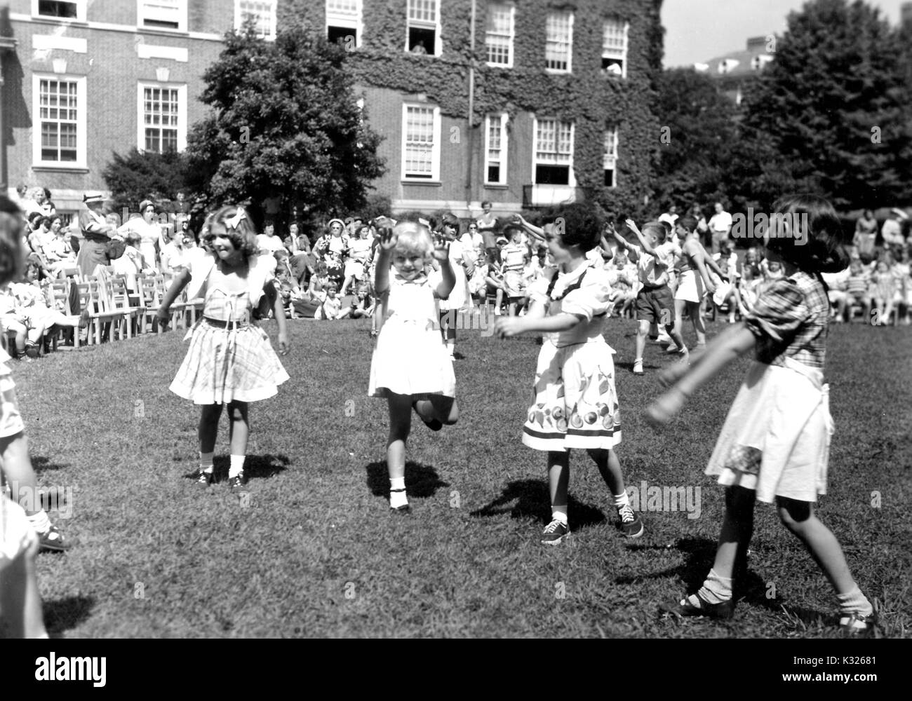 At the end of the school year for a demonstration school at Johns Hopkins University, young boys and girls put on a show in the grass on a sunny day, happily skipping and waving their hands in front of an audience made up of classmates, teachers, and parents sitting and standing outside of an ivy-covered campus building, Baltimore, Maryland, July, 1950. Stock Photo