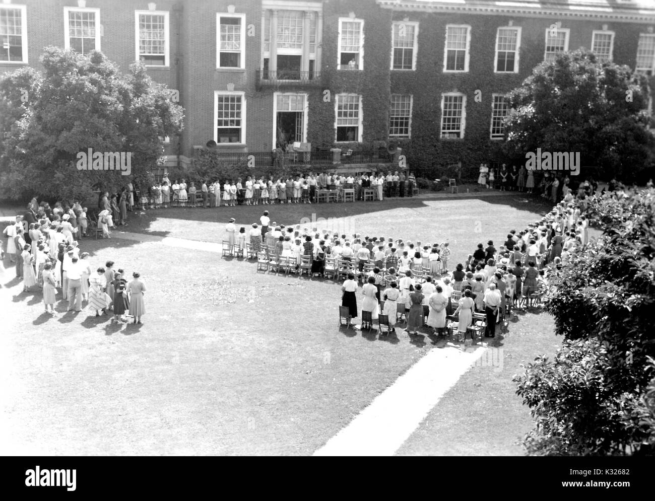 At the end of the school year for a demonstration school at Johns Hopkins University, parents and teachers stand in front of rows of chairs outside of an ivy-covered campus building, waiting for the young boys and girls to begin the show they will perform to close out the year, in the grass on the quadrangle on a sunny day, Baltimore, Maryland, July, 1950. Stock Photo
