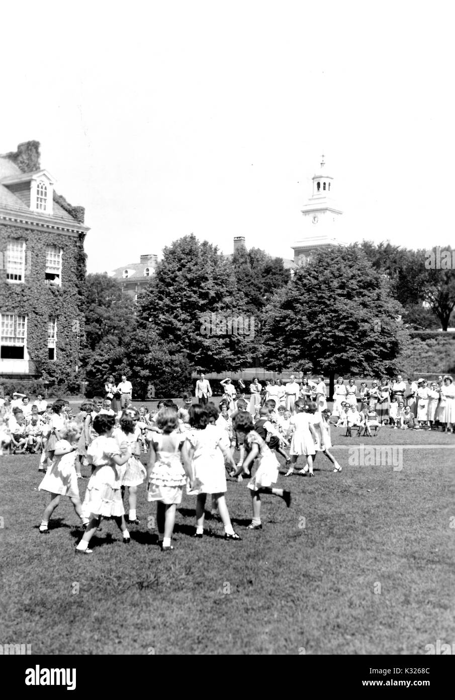 At the end of the school year for a demonstration school at Johns Hopkins University, young boys and girls put on a show in the grass on a sunny day, happily skipping dancing in front of an audience made up of classmates, teachers, and parents sitting and standing outside of an ivy-covered campus building, with Gilman Hall, a humanities building on campus, peeking through the trees in the distance, Baltimore, Maryland, July, 1950. Stock Photo